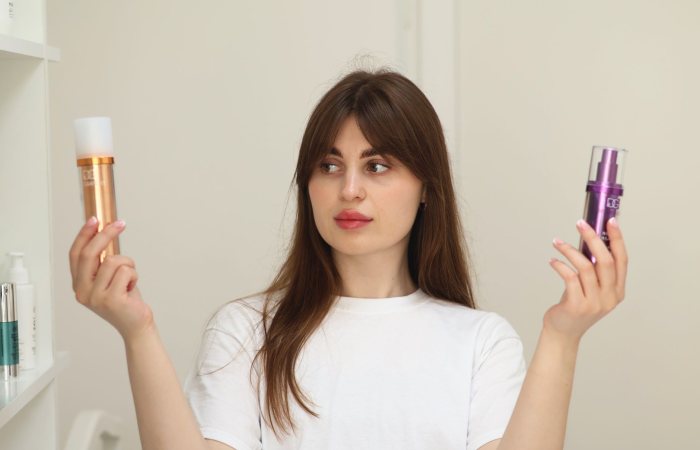 A young woman compares two cosmetic products while standing indoors, unsure of which to choose.
