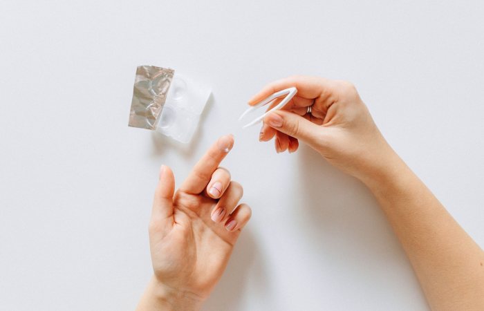 Hands holding a contact lens with tweezers next to a contact lens case on a white background.