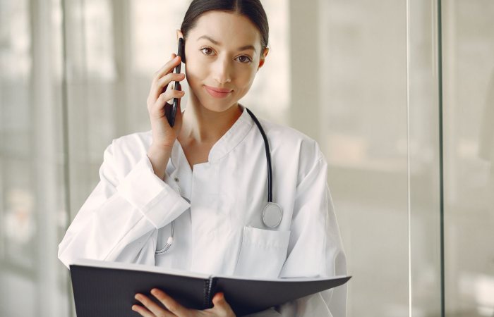 Crop ethnic female doctor in medical uniform with stethoscope and notebook talking on cellphone while standing in clinic corridor