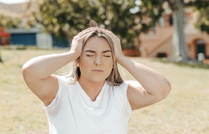 Adult woman holding her head in pain, sitting outdoors under sunlight, likely experiencing a migraine.