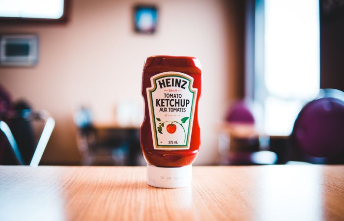 A Heinz Tomato Ketchup bottle on a wooden table with blurred background indoors.