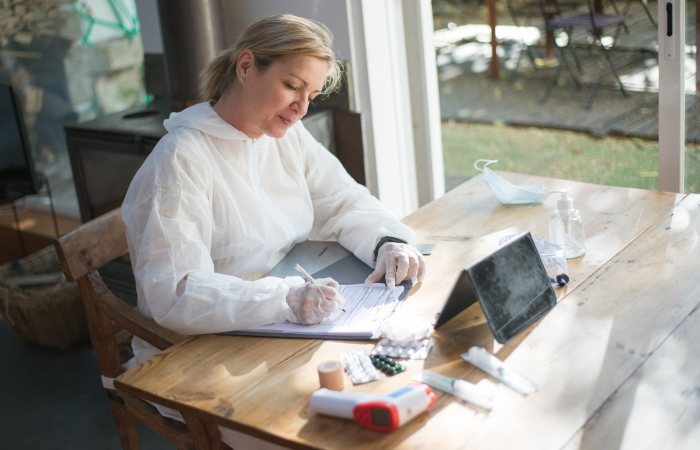 A healthcare worker in protective gear documents patient data indoors during daylight.