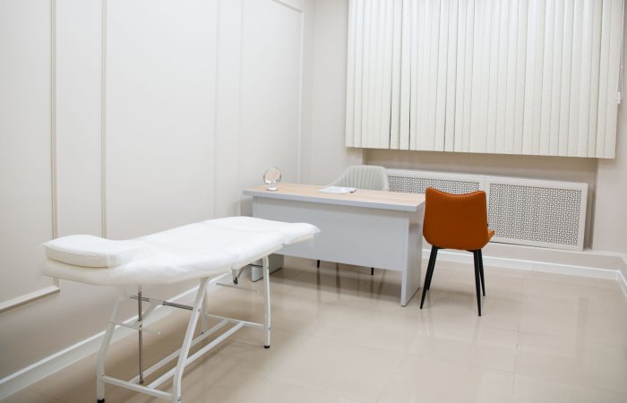 A minimalist medical examination room featuring a desk, chairs, and an examination bed in a sterile, neutral setting.